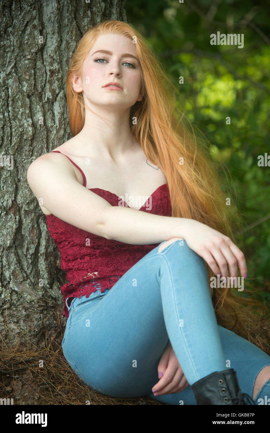 Belle tête rouge femme en skinny jeans et haut rouge, assis sur des aiguilles de pin avec en arrière contre un arbre. Banque D'Images