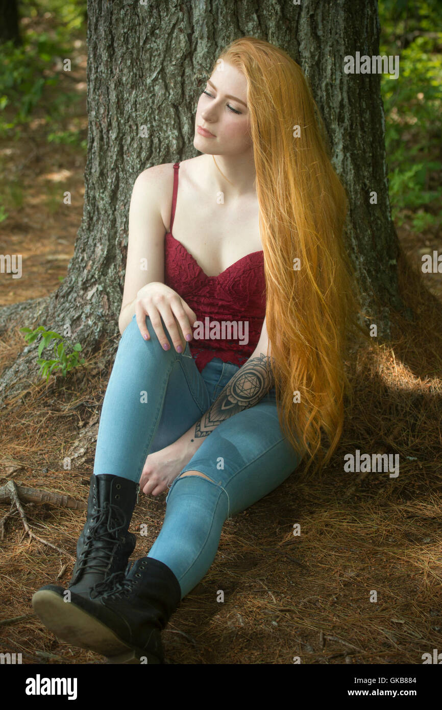 Belle tête rouge femme en skinny jeans et haut rouge, assis sur des aiguilles de pin avec en arrière contre un arbre. Banque D'Images