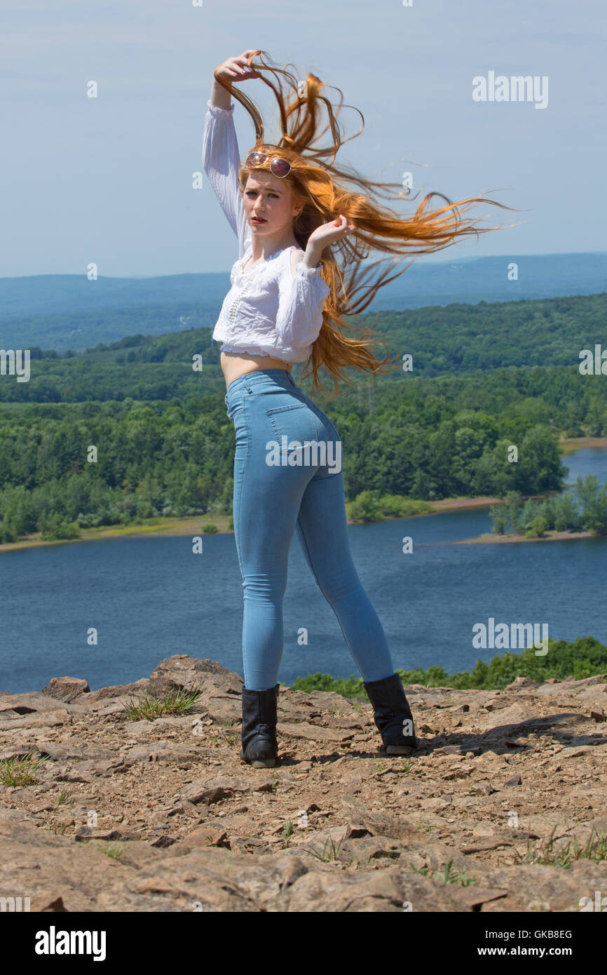 Belle tête rouge en skinny jeans, bottes noires, et haut blanc, en montagne avec le vent dans les cheveux et un lac ci-dessous. Banque D'Images