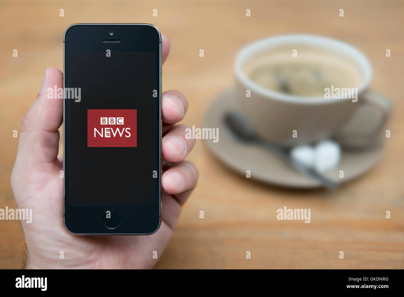 Un homme se penche sur son iPhone qui affiche le logo de BBC News, en restant assis avec une tasse de café (usage éditorial uniquement). Banque D'Images