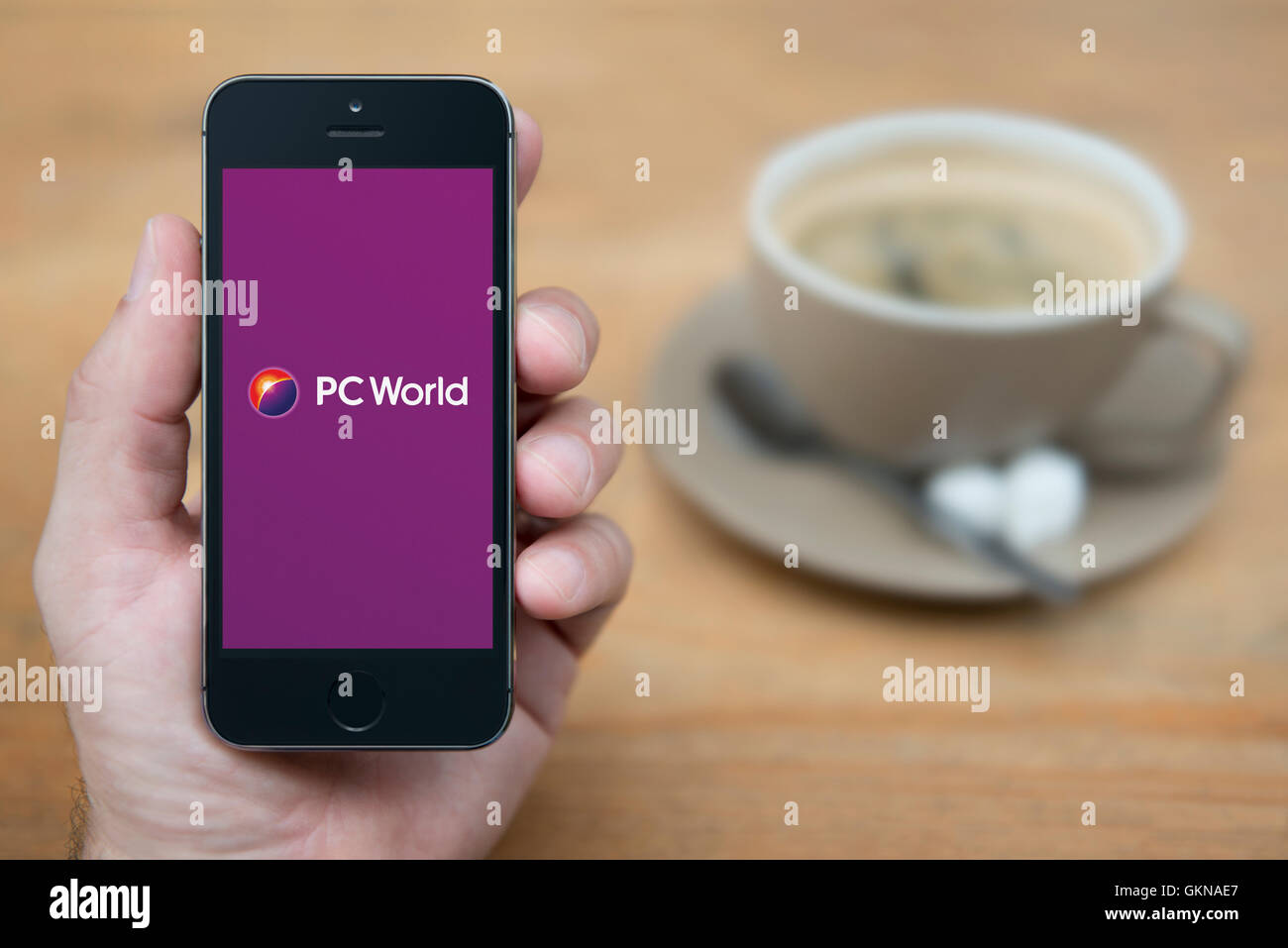Un homme se penche sur son iPhone qui affiche le logo PC World, alors qu'assis avec une tasse de café (usage éditorial uniquement). Banque D'Images