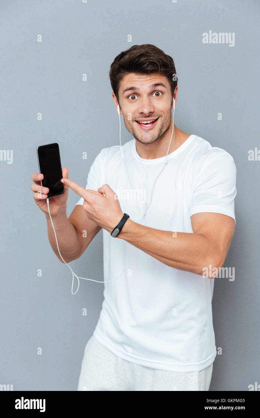 Cheerful Young man with earphones doigt sur smartphone Avec écran blanc sur fond gris Banque D'Images
