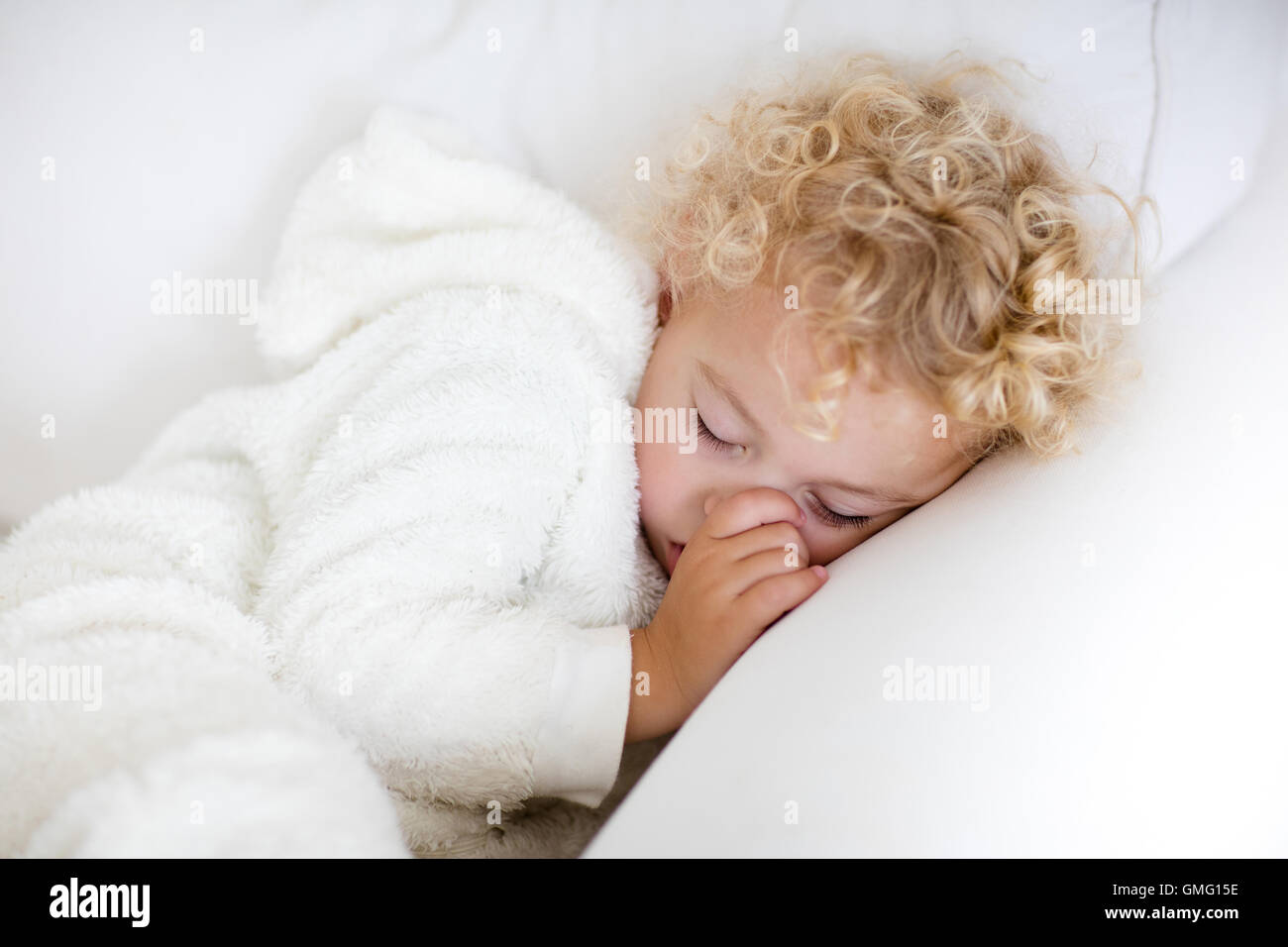 Cute blonde curly petit garçon endormi sur la table blanche. Tout-petits fatigués prendre une sieste portant des pyjamas blanc chaud, suçant son pouce. Banque D'Images