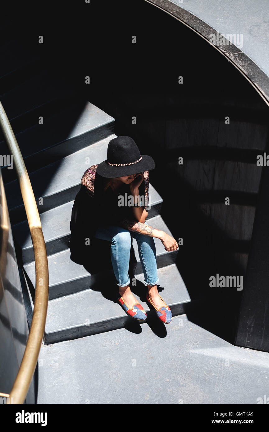 Fille aux tatouages en vêtement élégant et hat assis dans les marches Banque D'Images