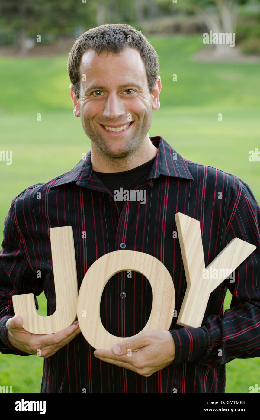 Jeune homme tenant les lettres du mot 'JOY' à l'extérieur portant une chemise rouge et noir. Banque D'Images