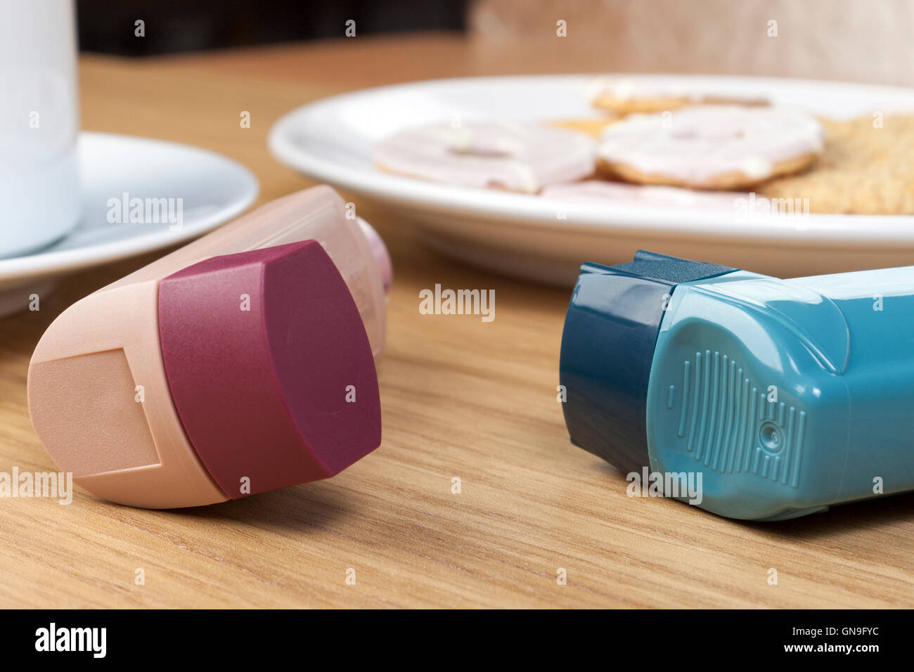 Gros plan d'un asthme inhalateur brun et bleu sur la table du petit déjeuner avec une tasse de thé et une assiette de biscuits Banque D'Images