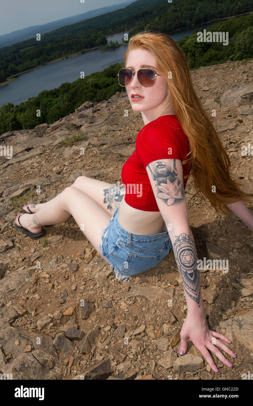Belle femme avec de longs cheveux rouges, en jeans et chemise rouge, assis sur les rochers de la montagne en été, avec le lac. Banque D'Images