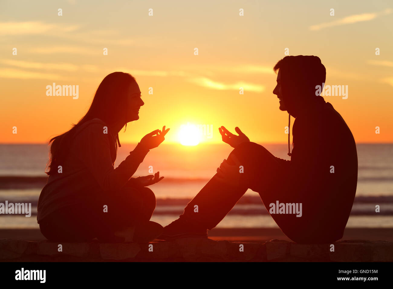 Vue latérale d'un corps complet de deux amis ou un couple silhouette d'adolescents assis et parler au lever du soleil sur la plage Banque D'Images