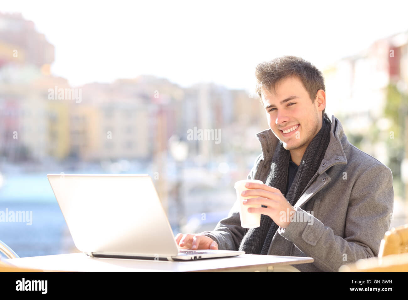 Beau et élégant, l'homme à l'aide d'un ordinateur portable et à la recherche à l'appareil photo dans un café terrasse en hiver dans un port de l'urbanisation Banque D'Images