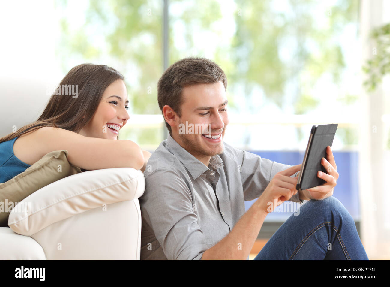 Cheerful couple using a tablet à la ligne assis dans le salon à la maison avec une fenêtre à l'arrière-plan Banque D'Images