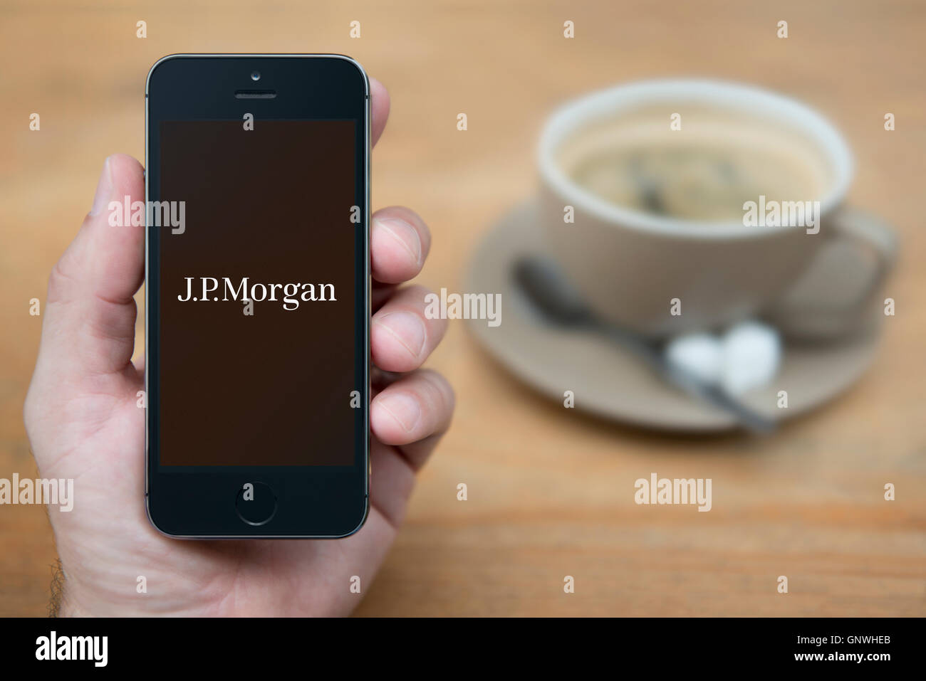 Un homme se penche sur son iPhone qui affiche le logo de J. P. Morgan Bank, en restant assis avec une tasse de café (usage éditorial uniquement). Banque D'Images