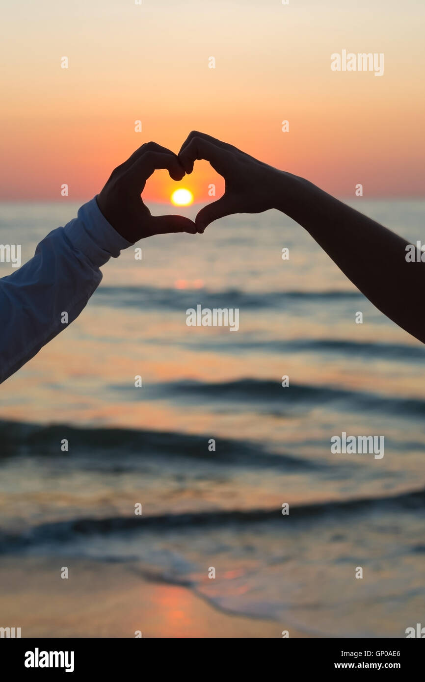 Couple forme de cœur avec leurs mains sur une plage, au coucher du soleil. Banque D'Images