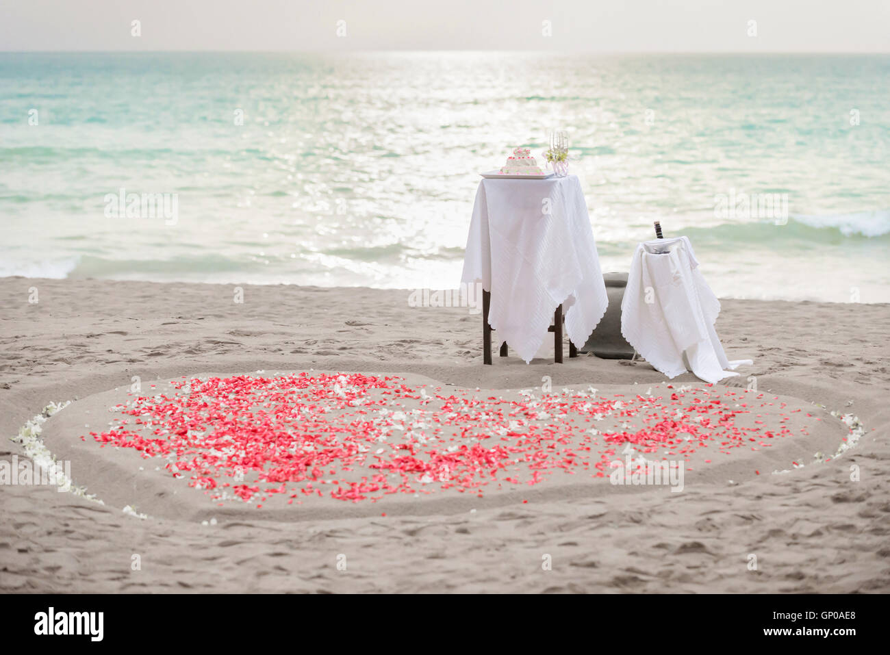 Table pour la cérémonie du mariage, le gâteau, le champagne et hearth forme des pétales blanc et rouge sur le sable, le coucher du soleil. Copier l'espace. Banque D'Images