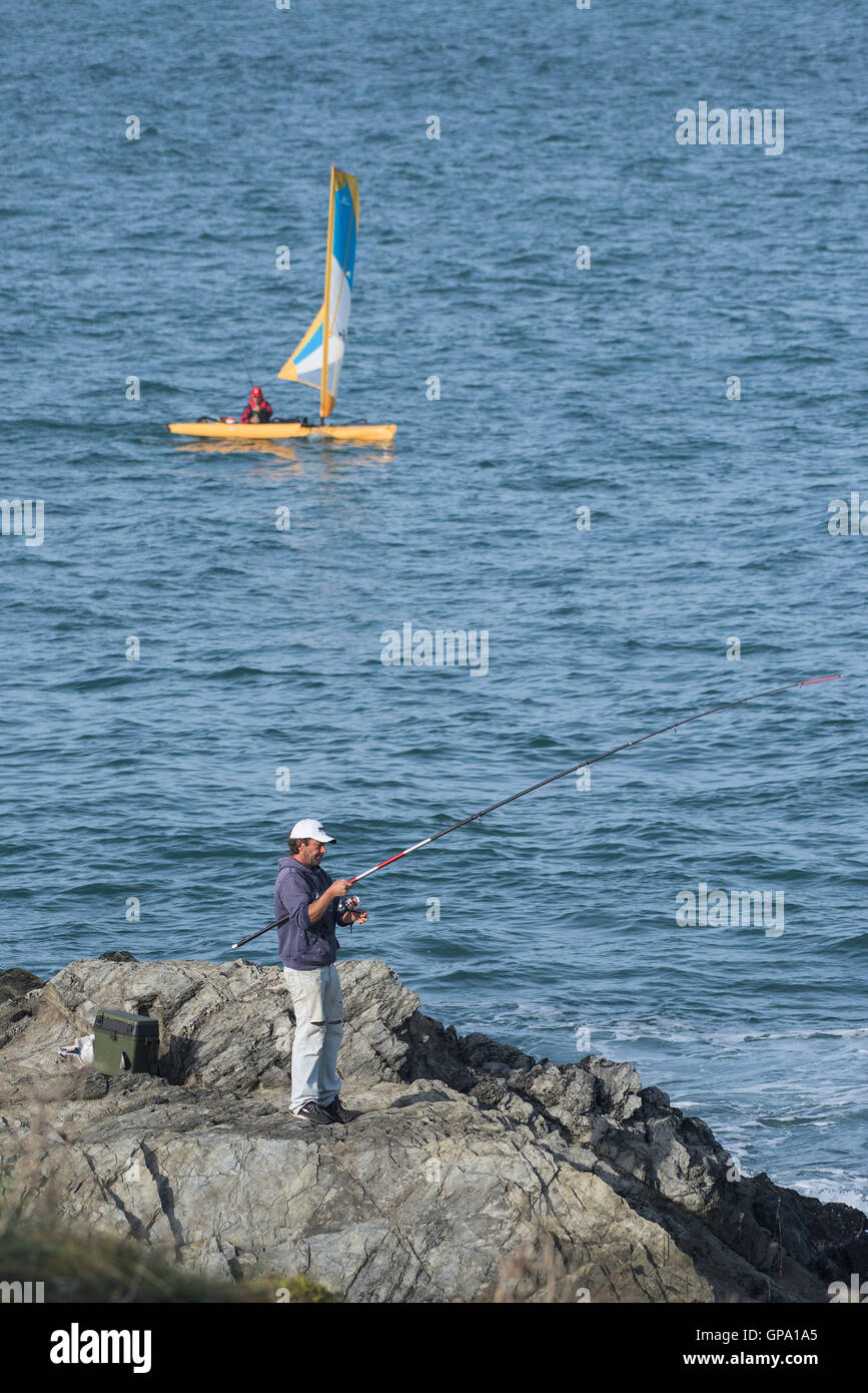 Un homme poissons de rochers comme un trimaran jaune sails passé en arrière-plan. Banque D'Images