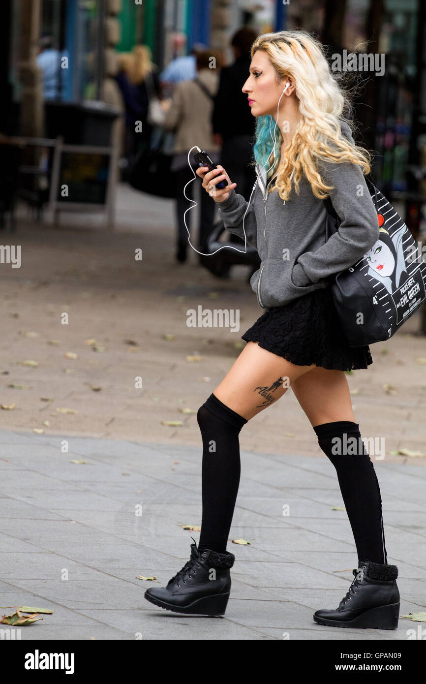Une grande femme avec de longs cheveux blonds wearing earphones écoute de la musique sur son téléphone mobile en marchant à Dundee, Royaume-Uni Banque D'Images