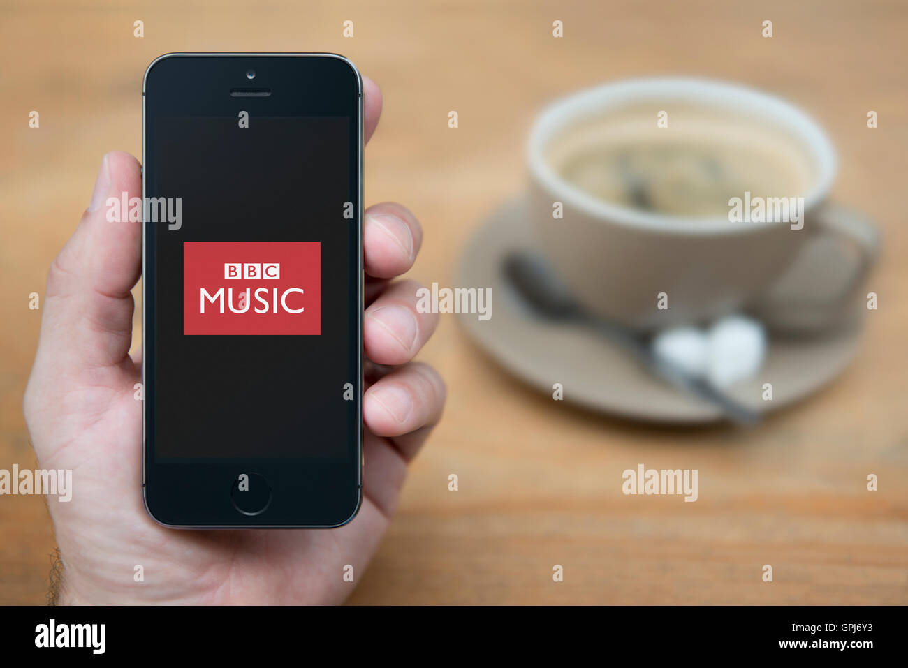 Un homme se penche sur son iPhone qui affiche le logo de BBC Music, tandis qu'assis avec une tasse de café (usage éditorial uniquement). Banque D'Images