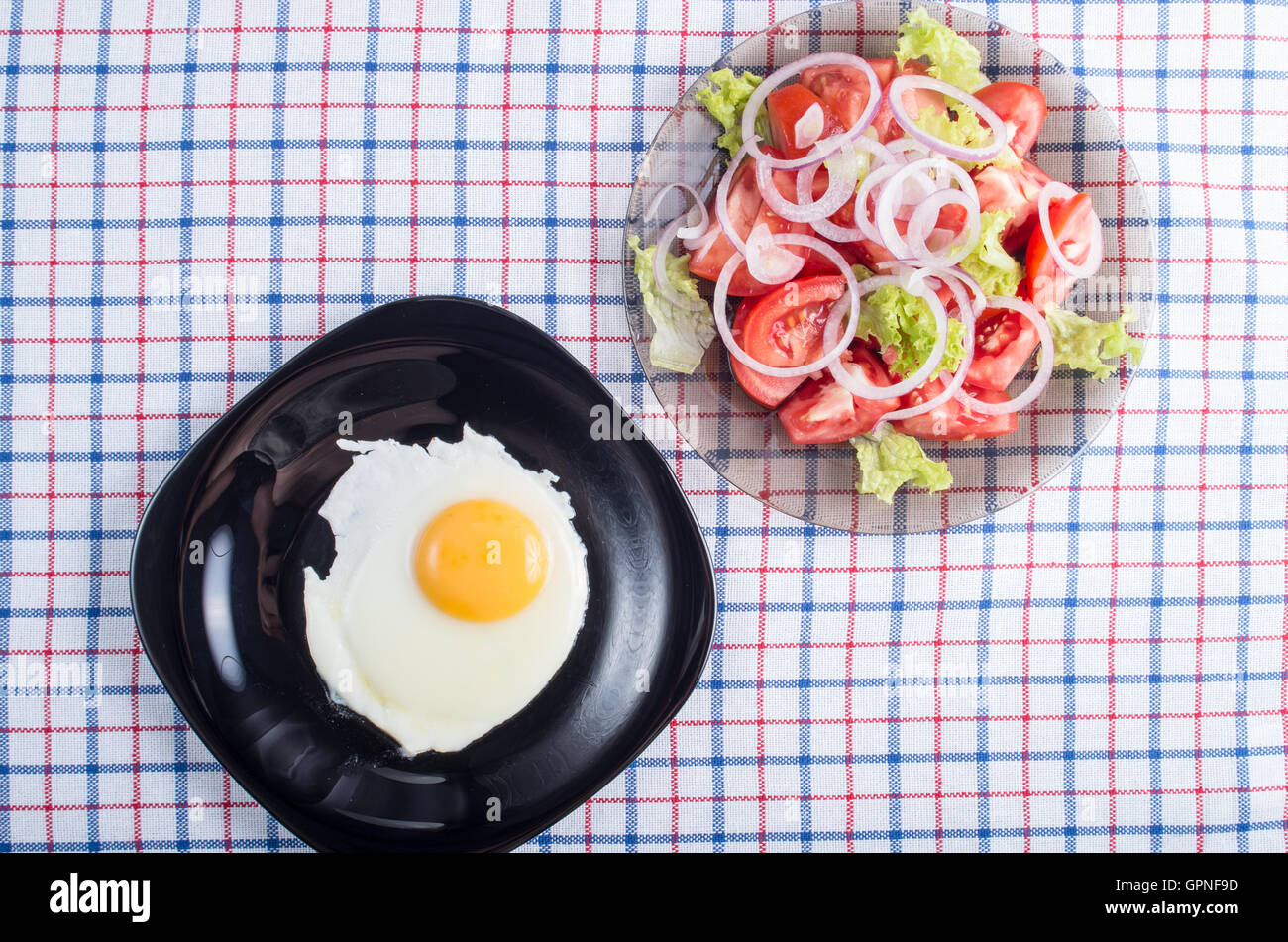 Vue de dessus de la surface de la table avec une plaque noire avec un œuf frit et une petite portion de Salade de tomates sur un tissu tablecl Banque D'Images