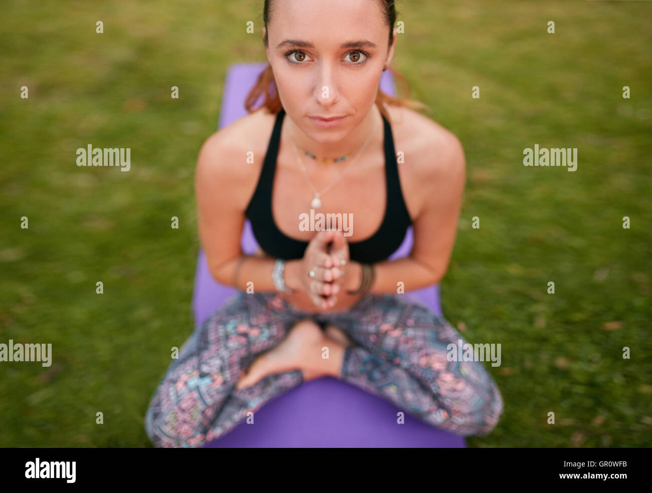 Vue de dessus de la femme de fitness assise sur le tapis d'exercice avec ses mains jointes et regardant la caméra. femme de fitness faisant du yoga à l'extérieur Banque D'Images