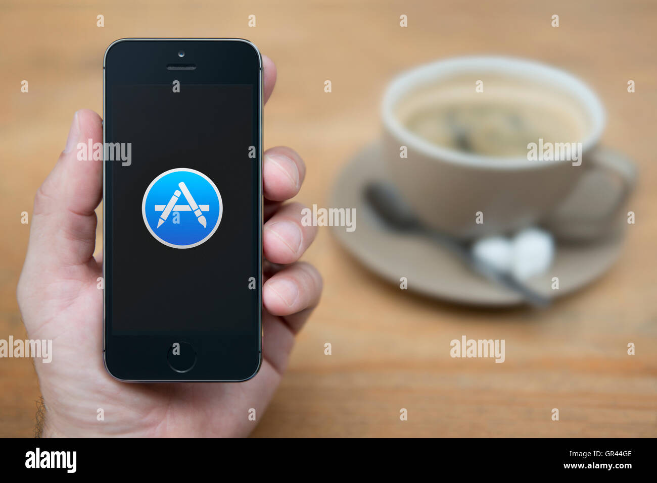 Un homme se penche sur son iPhone qui affiche le logo Apple App Store, tandis que sam avec une tasse de café (usage éditorial uniquement). Banque D'Images