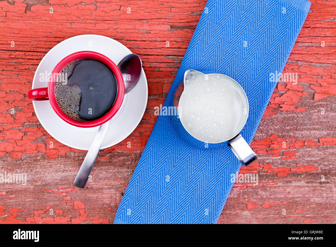 Fraîchement préparée et tasse de café noir dans une tasse rouge sur une soucoupe blanc servi avec un pichet de lait et sur une serviette bleu grunge r en bois Banque D'Images