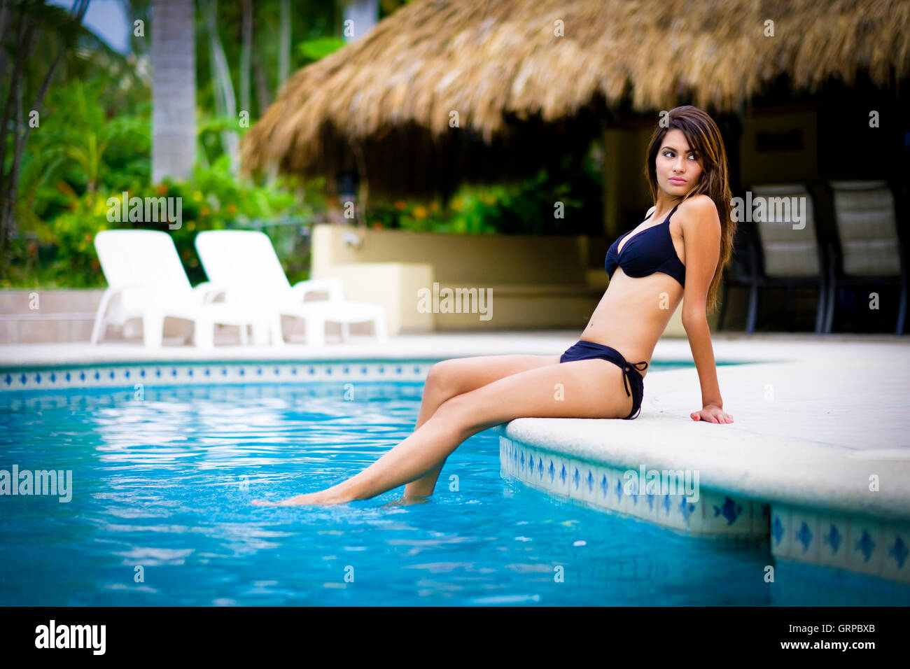 Young attractive latina woman in bikini profitant à siéger à une piscine résidentielle palapa avec en arrière-plan. Banque D'Images
