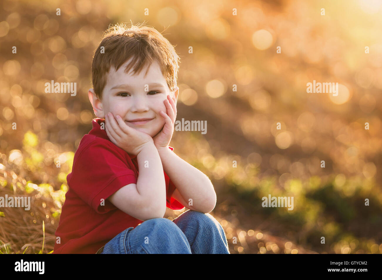 Un mignon jeune garçon sourit à la caméra, tenant sa tête dans ses mains, avec un arrière-plan flou d'Effervescents. Banque D'Images