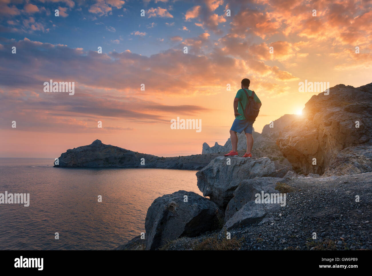 Jeune homme debout avec sac à dos à la pierre sur le bord de la mer au ciel  coucher de soleil coloré. Beau paysage avec homme sportif, des rochers  Photo Stock -