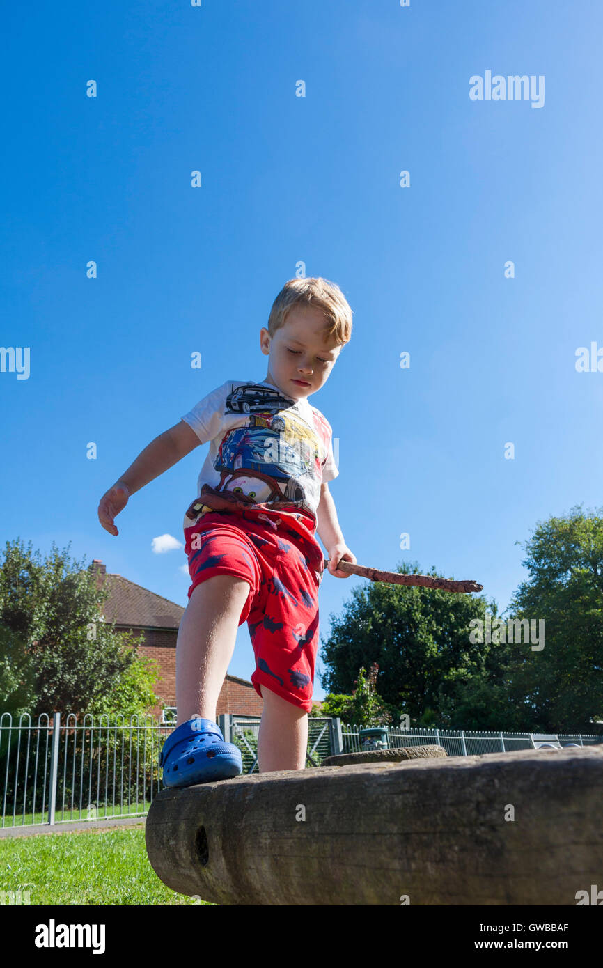 Un young caucasian boy holding a stick marche sur la poutre dans un parc. Ickenham, London, UK Banque D'Images