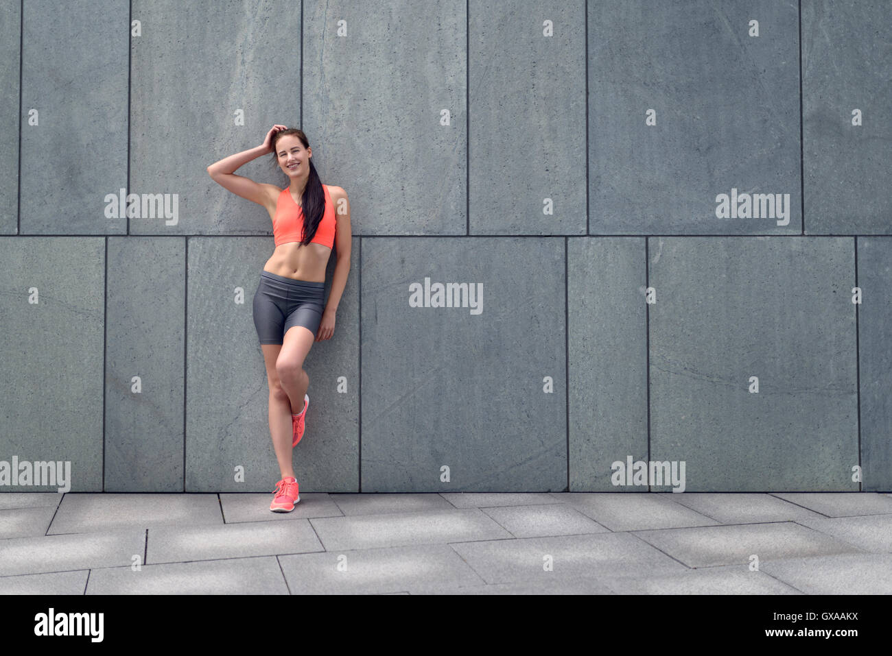 Jolie jeune femme sportive s'appuyant sur un mur carrelé gris d'un bâtiment urbain avec sa main sur ses longs cheveux bruns et un lovel Banque D'Images