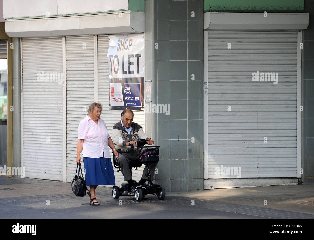 Une vue générale de femme marche à côté d'un homme à l'aide d'un scooter d'invalidité, lorsqu'ils passent d'un magasin vide en entrée est, Harlow, Essex. Banque D'Images