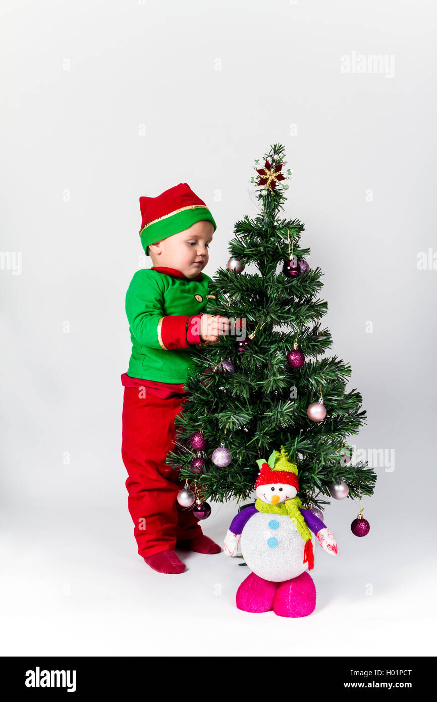 Baby Boy dressed as Santa's helper decorating Christmas Tree, hanging ornaments. fond blanc. Banque D'Images