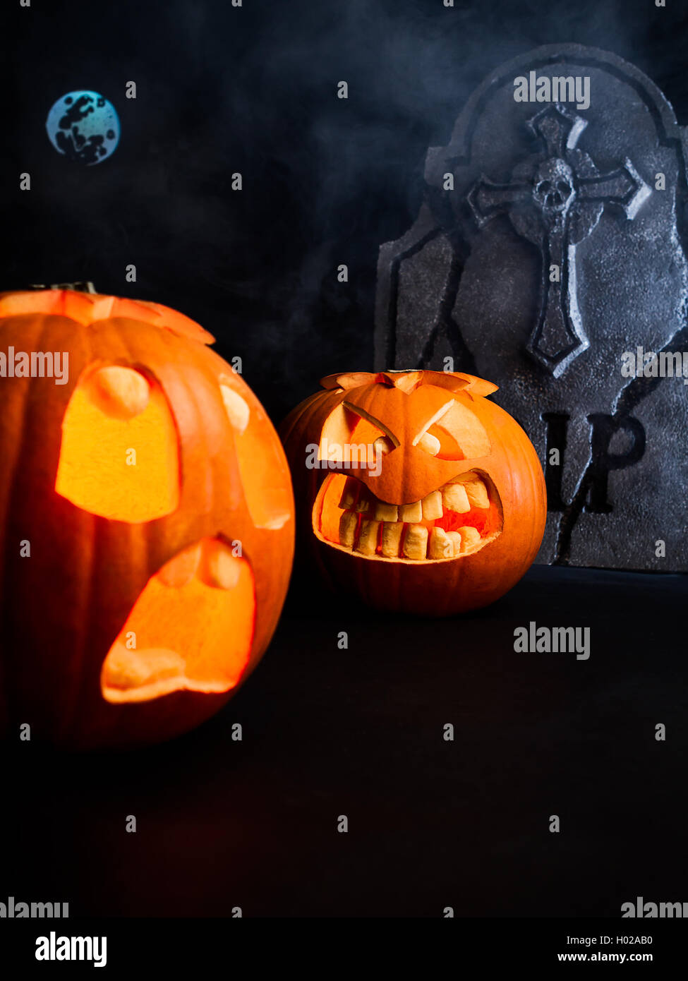 Peur et visage en colère face à des citrouilles d'halloween avec lune et pierre tombale sur misty fond sombre. Banque D'Images