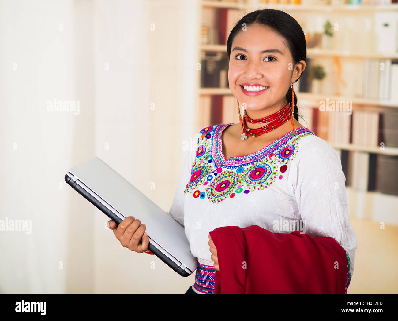 Beau jeune avocat portant blouse traditionnelle des Andes avec collier, holding laptop et red jacket smiling, étagères backg Banque D'Images