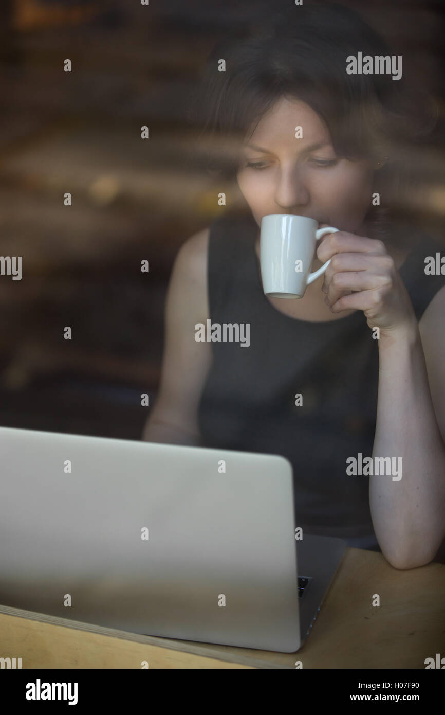 Portrait de belle jeune femme assise dans la tasse de café moderne verre, sirotant du café, working on laptop Banque D'Images