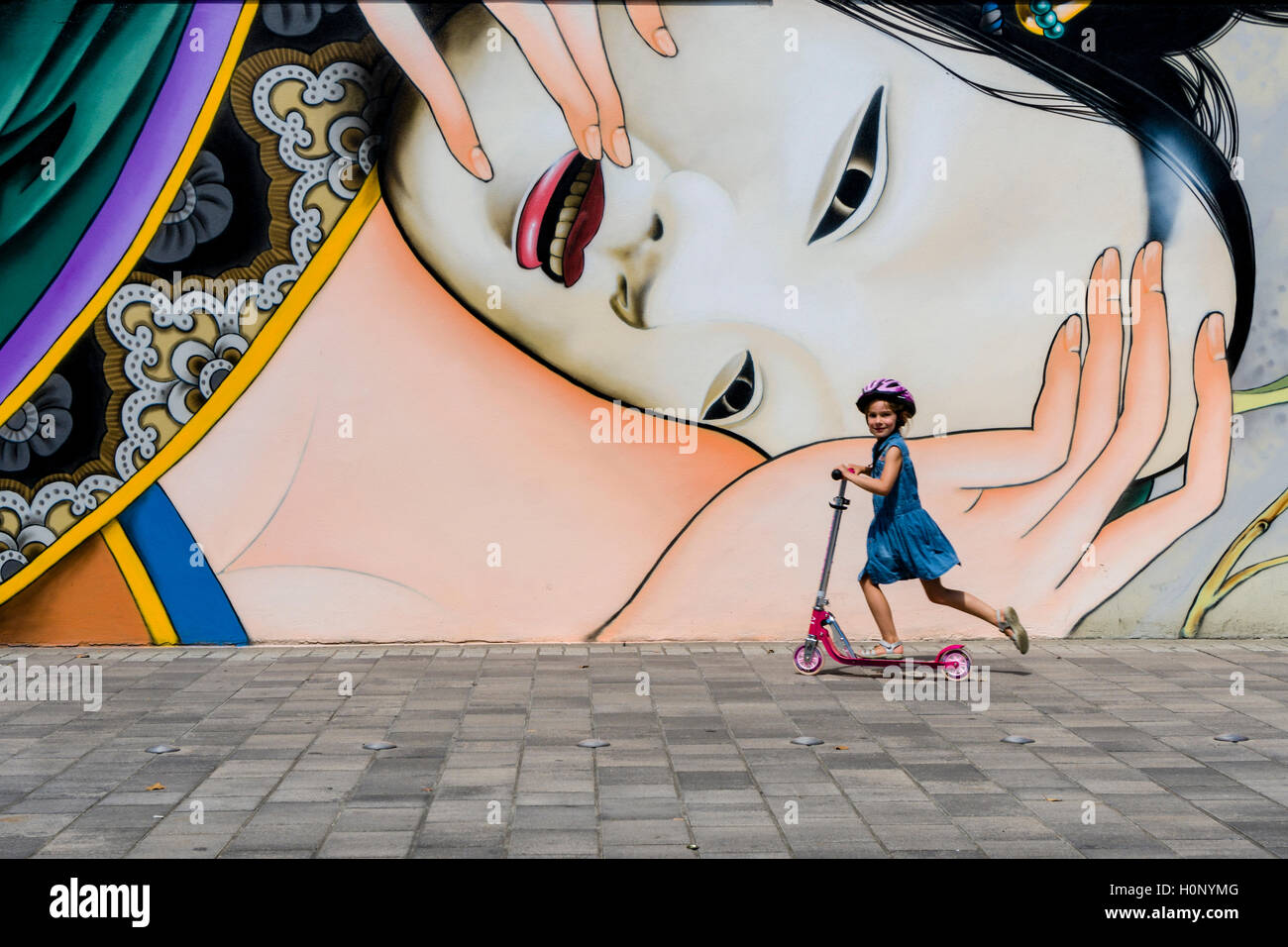Un graffity avec motif japonais est peint dans un mur de la maison, une petite fille passe sur un scooter pour enfants Banque D'Images