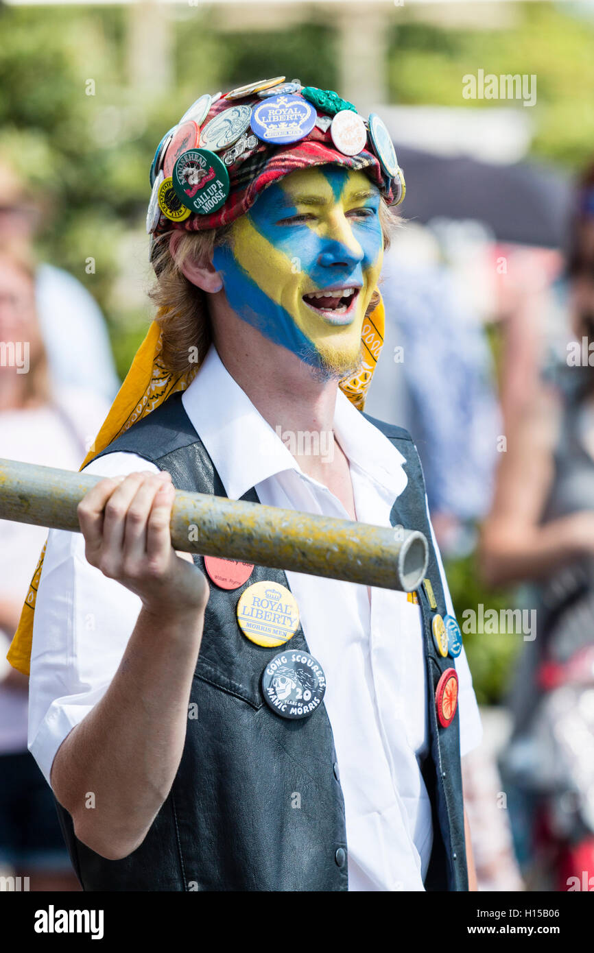 Close up of Royal Liberty Morris dancer, jeune homme, le visage peint en jaune et bleu, porte bouchon rouge couvert de badges, l'escrime avec poteau de métal. Banque D'Images