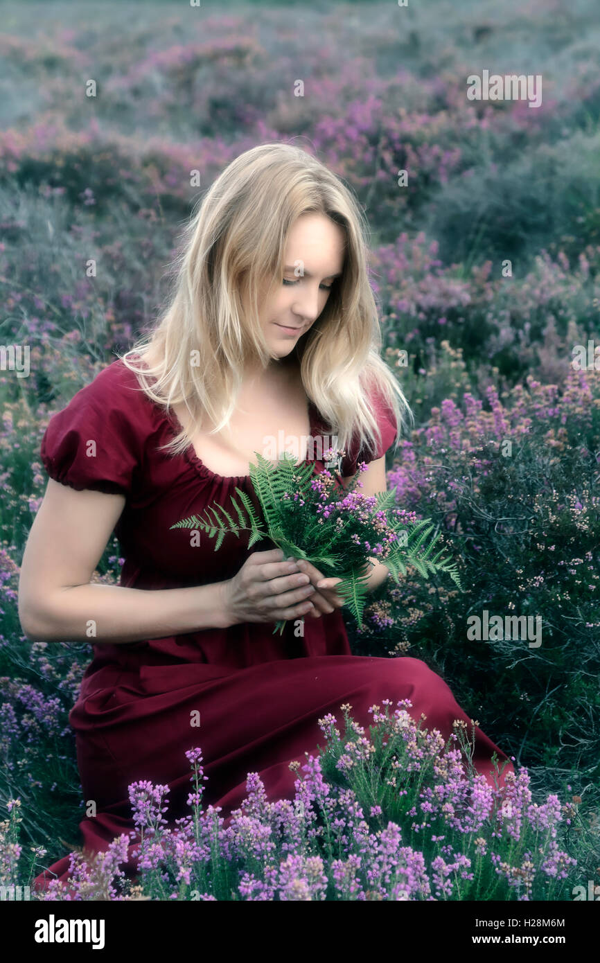 Une femme en robe rouge est assise dans la bruyère avec un bouquet de fleurs Banque D'Images