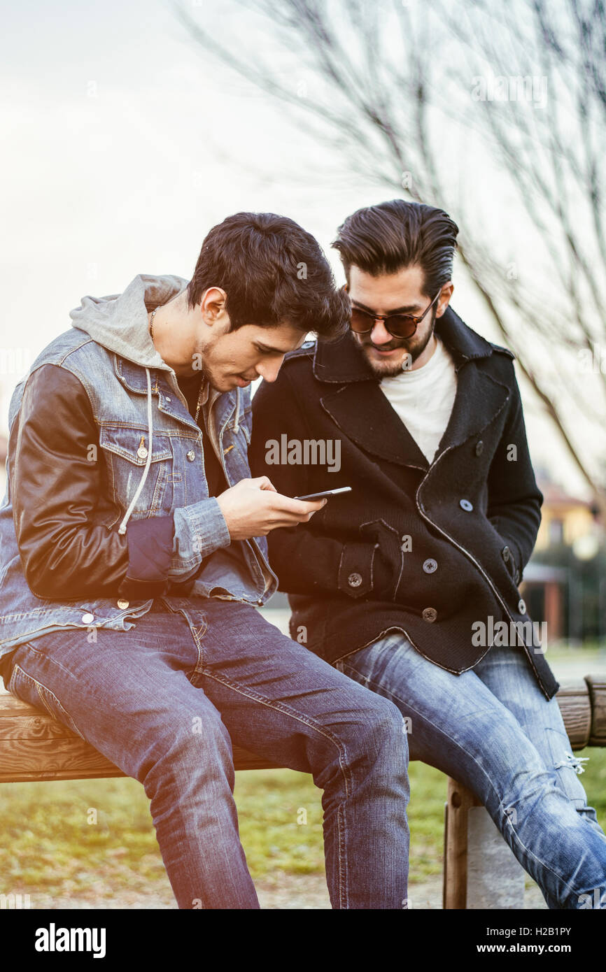Deux jeunes hommes casual mode assis ensemble sur un banc à l'extérieur de la lecture d'un SMS ou message texte, ou regarder des photos sur un mobil Banque D'Images