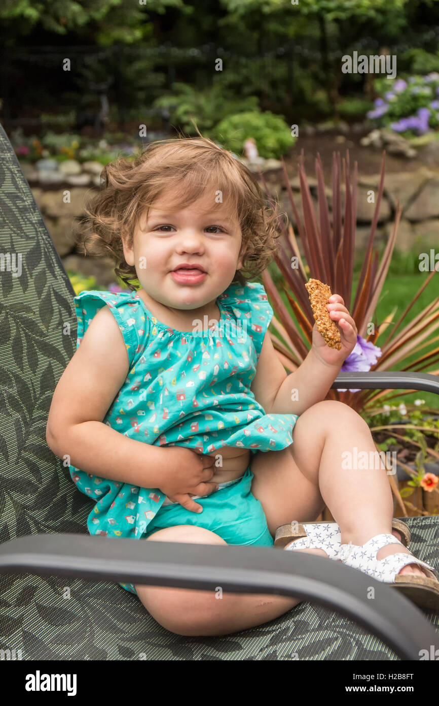 Dix-huit mois, fille, assise dans une chaise de patio à l'extérieur manger une barre granola, tapotant son ventre, à Issaquah, Washington, USA Banque D'Images