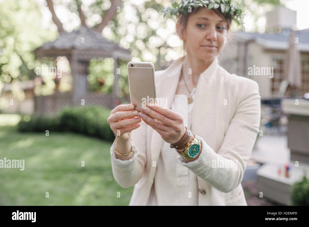 Femme avec une couronne de fleurs dans les cheveux, debout dans un jardin, en tenant un téléphone mobile selfies avec elle. Banque D'Images