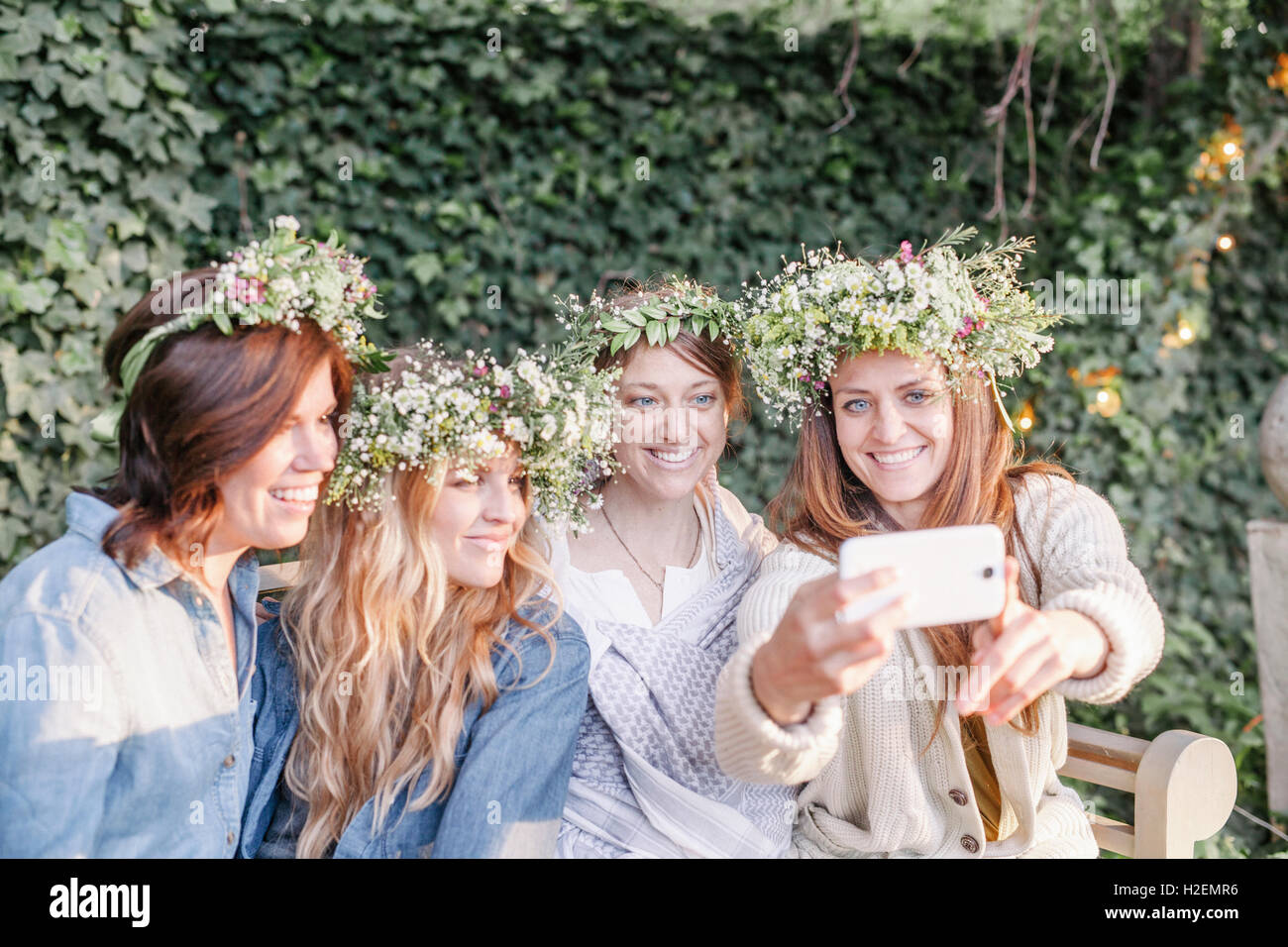 Quatre femmes avec des couronnes de fleurs souriant dans leurs cheveux assis dans un jardin, en tenant un. selfies Banque D'Images