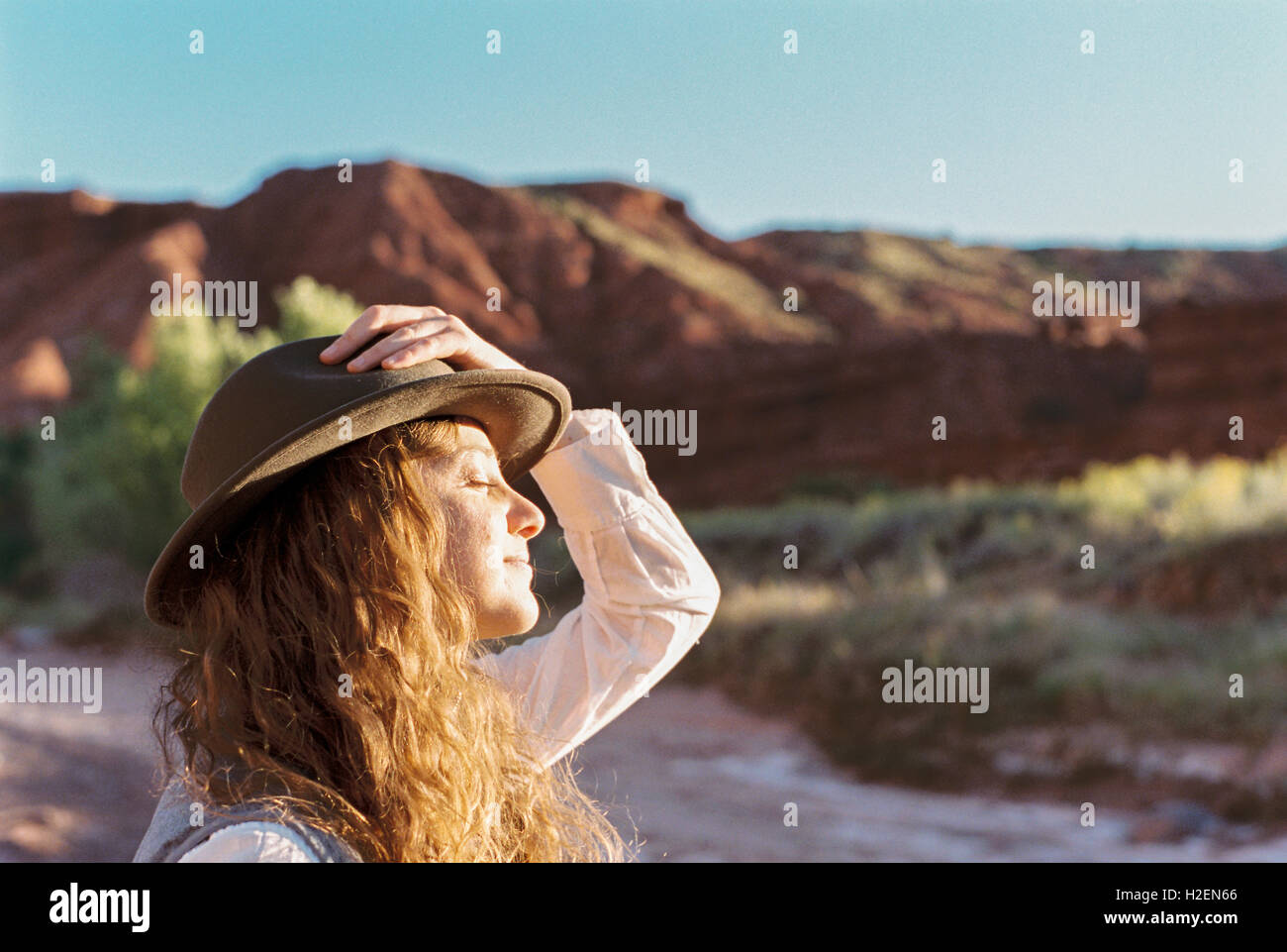 Une femme portant un chapeau debout dans un espace ouvert avec des montagnes, le visage vers le soleil. Banque D'Images