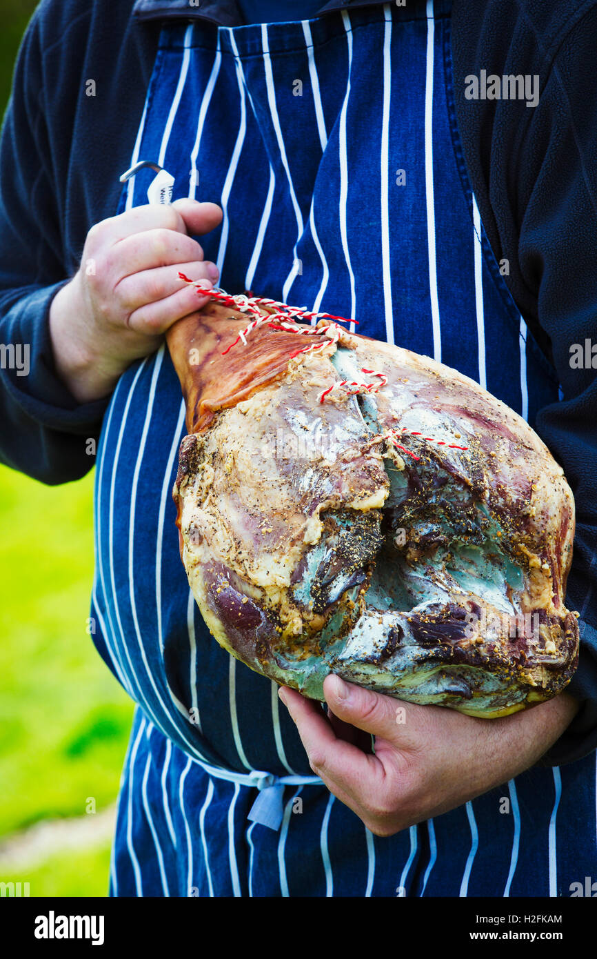 Boucher le port d'un tablier bleu à rayures , debout à l'extérieur, tenant un grand jambon séché. Banque D'Images