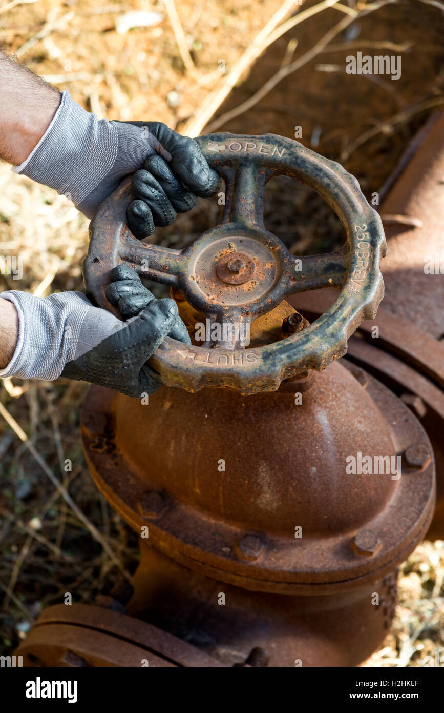 Gants de travail avec les mains serrant une valve du tuyau d'une très ancienne et ligne d'eau rouillée Banque D'Images