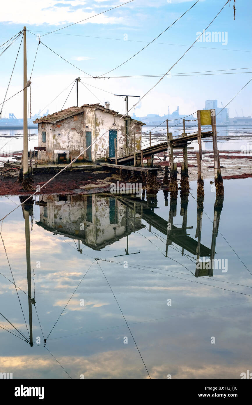 Ancienne maison de pêcheurs se reflétant dans la lagune de Marina di Ravenna, port industriel sur l'arrière-plan. Banque D'Images