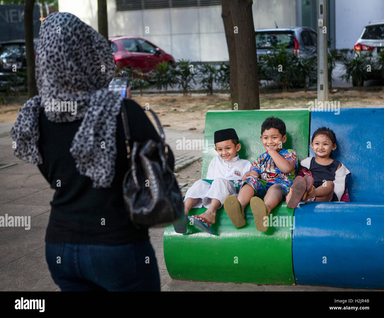 Heureux les enfants musulmans s'asseoir sur un banc de parc pendant que leur mère prend leur photo à Kuala Lumpur, Malaisie. Banque D'Images