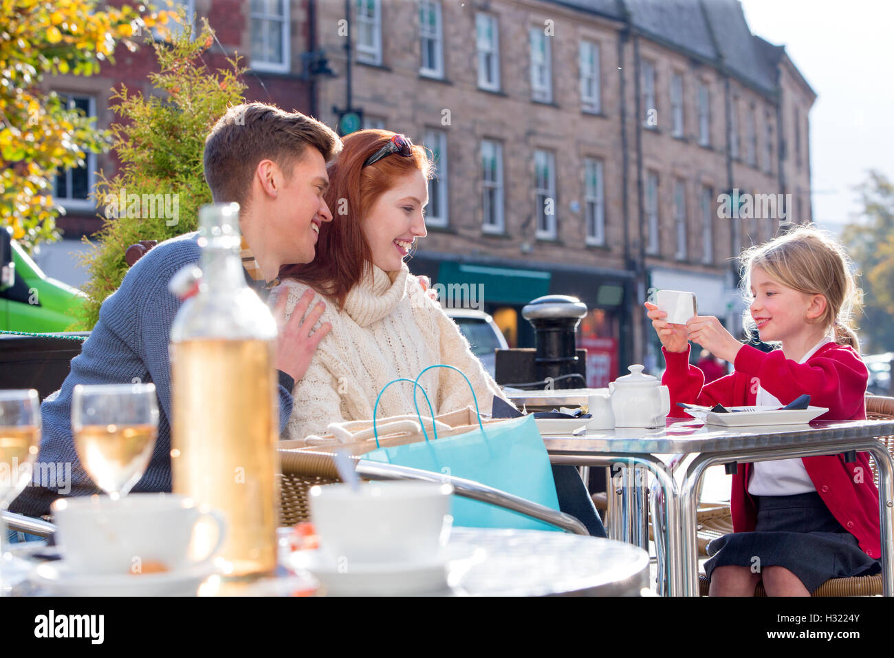 Petite fille de prendre une photo de ses parents sur un smartphone. Ils sont assis à l'extérieur, dans un café et la petite fille porte il Banque D'Images