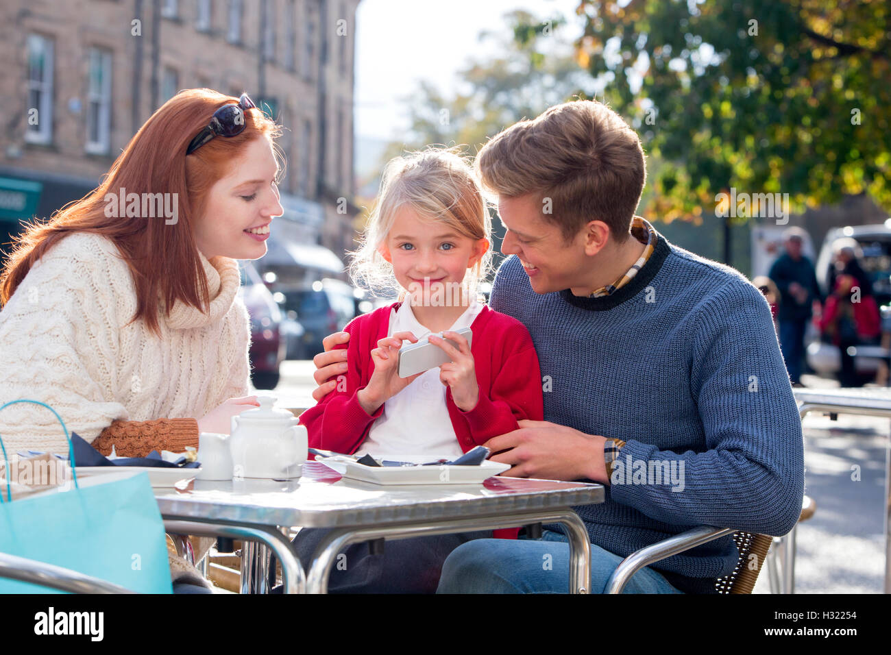 Famille de trois jeunes assis à l'extérieur, dans un café. La petite fille est maintenant un smartphone et regardant la caméra. Les parents Banque D'Images
