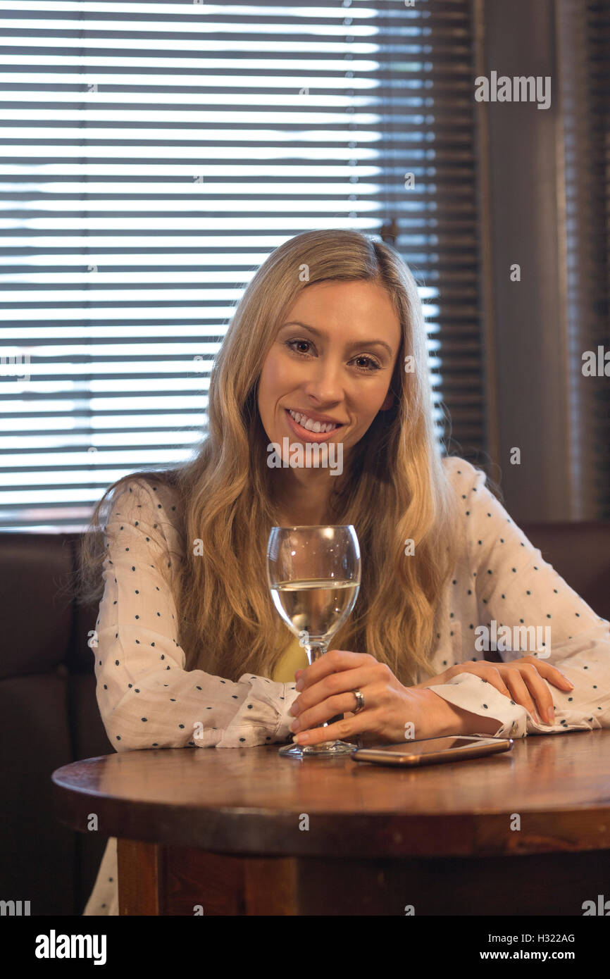 Belle jeune femme assise dans un pub avec un verre de vin et de son smartphone. Elle sourit à la caméra. Banque D'Images