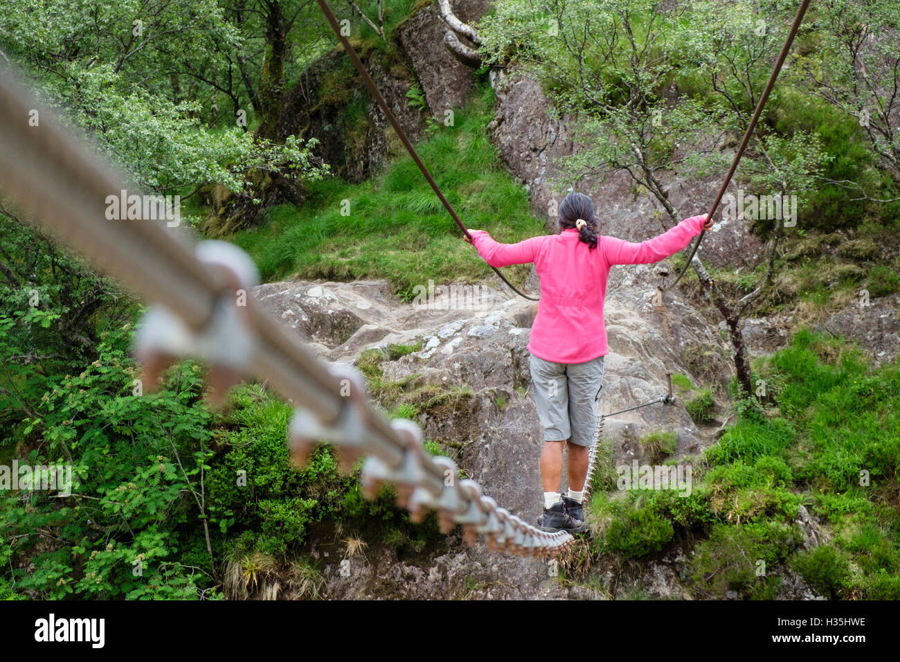 Asian Woman in pink marchant sur Steall wire rope bridge crossing Water de Nevis river. Fort William Glen Nevis Highland Ecosse Royaume-Uni Grande-Bretagne Banque D'Images
